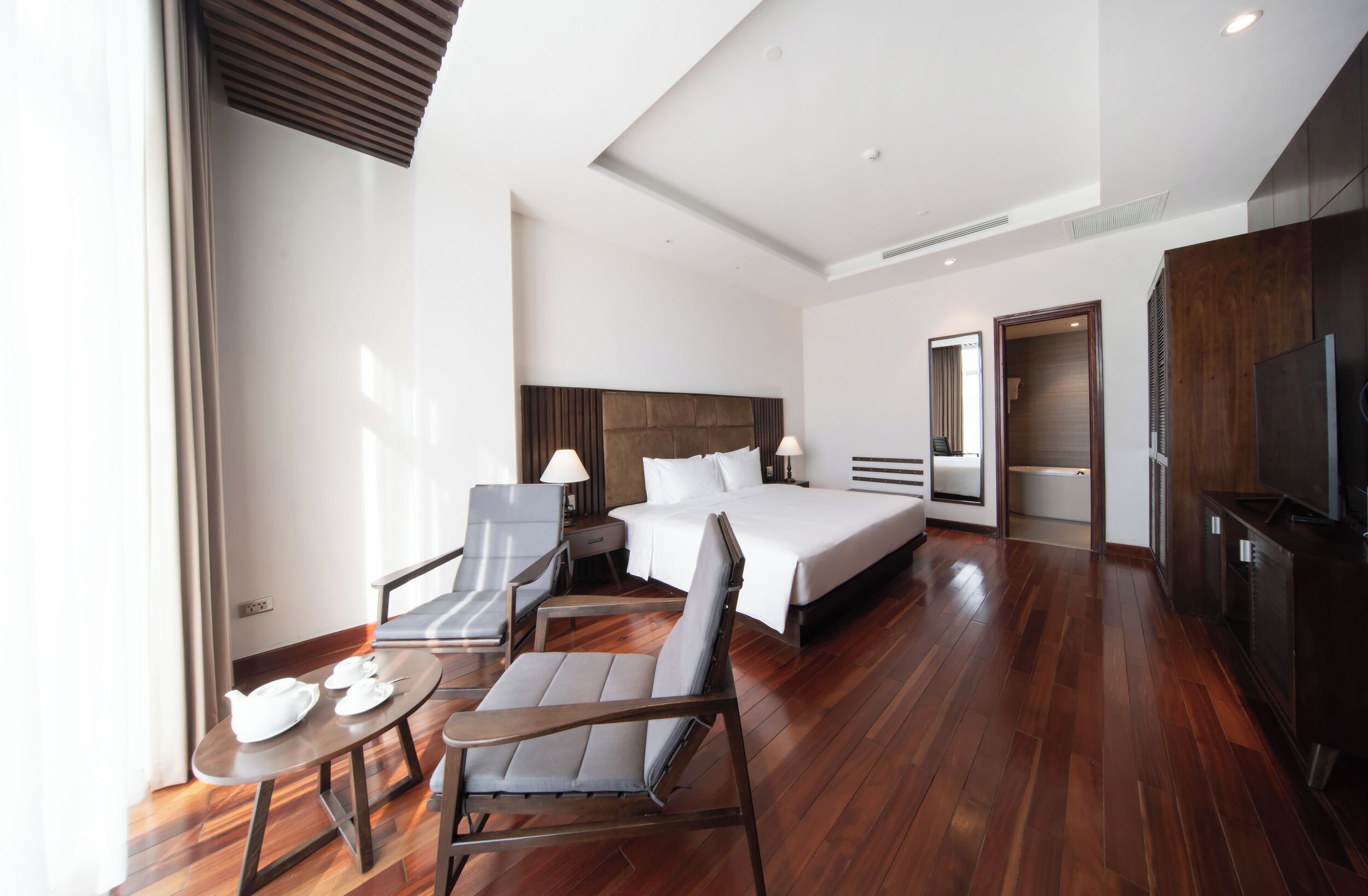 Quarto de hotel com uma cama grande, duas poltronas, uma mesa pequena com chá, uma televisão e piso de madeira.