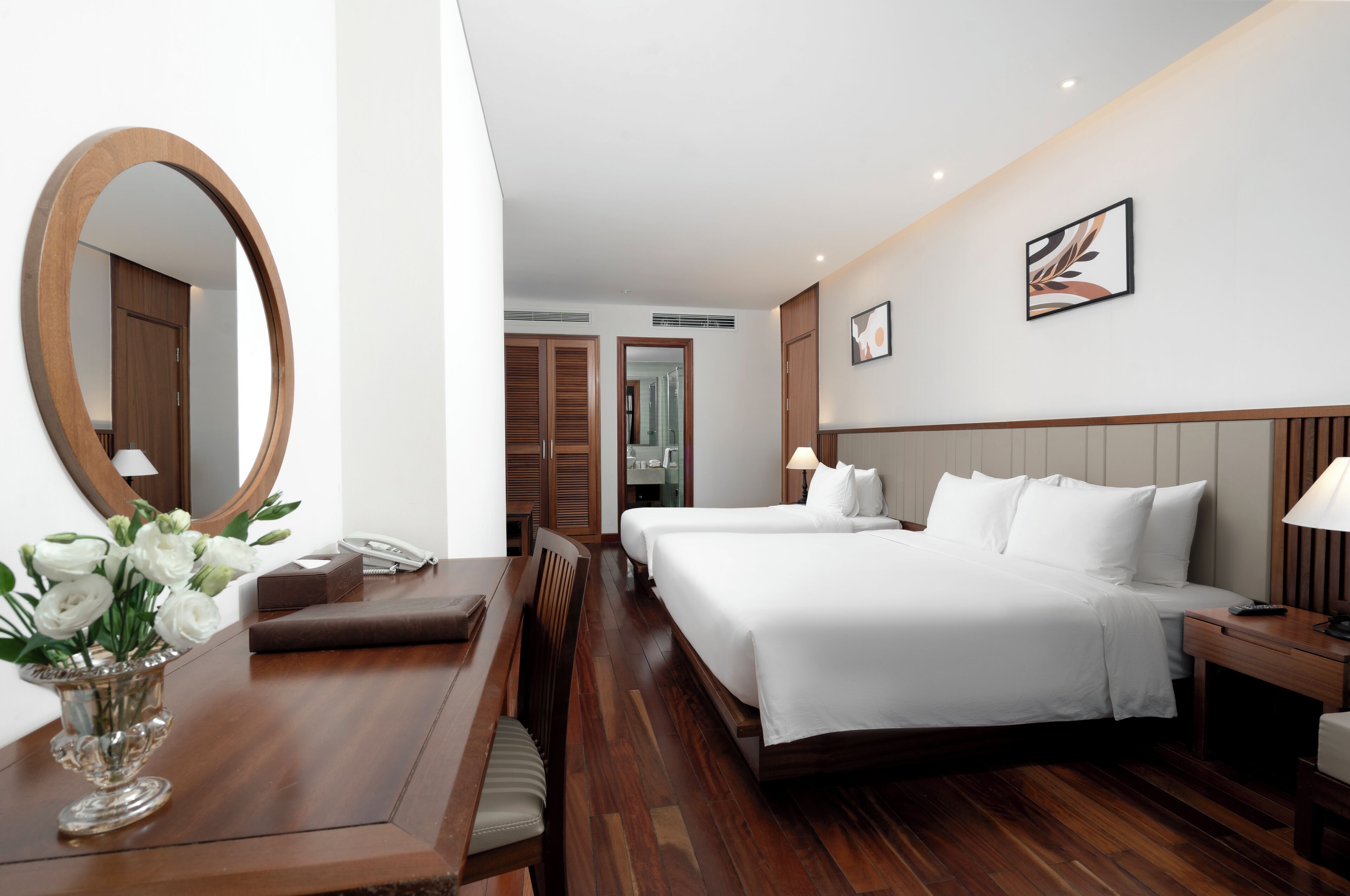 A hotel room with a large round mirror, a wooden desk with a vase of flowers, two beds with white linens, and a bedside table with a lamp.