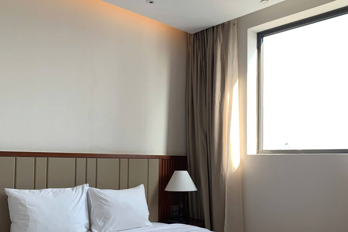 A hotel room with a large bed, a bedside lamp, a window with curtains, and a plain wall.