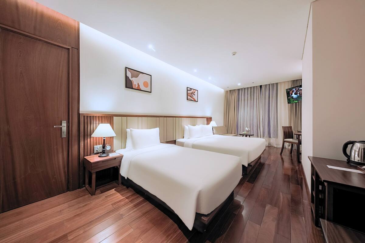 A hotel room with two beds, a wooden floor, a TV, and a desk with a kettle.