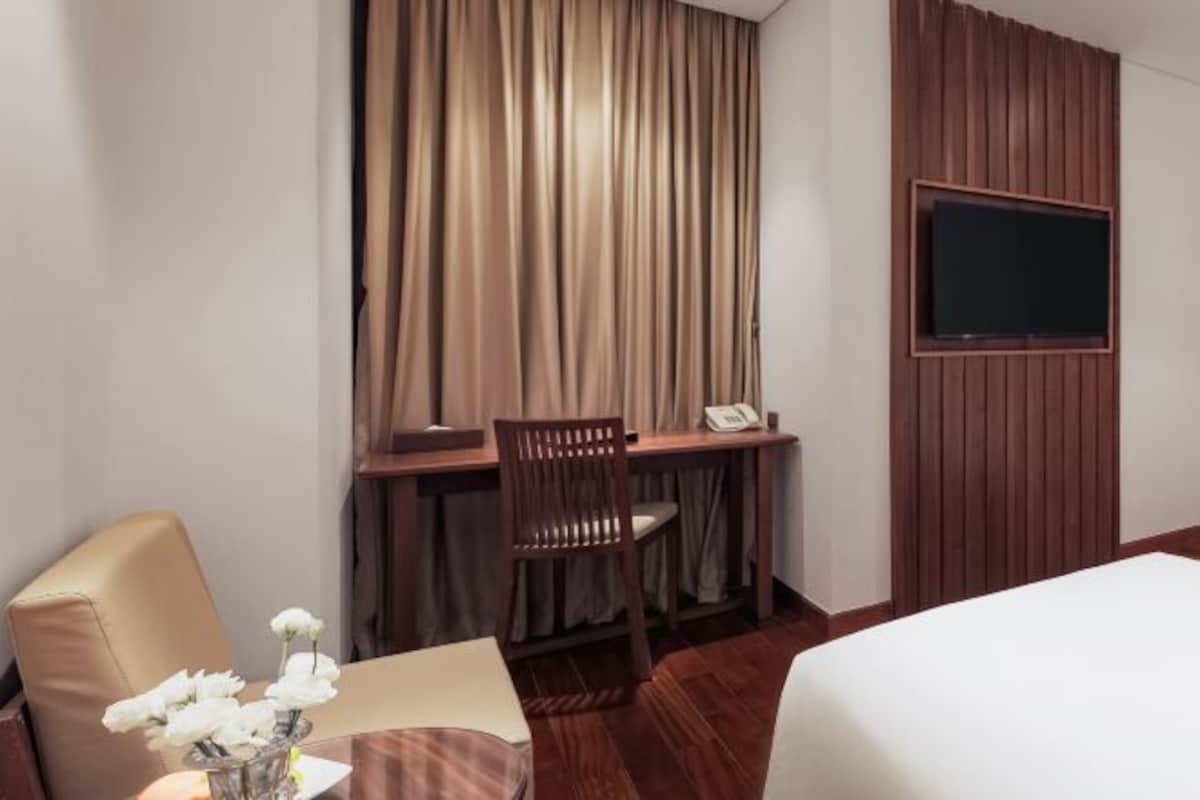 A hotel room with a bed, a desk with a chair, a TV mounted on the wall, and a vase with flowers on the table.