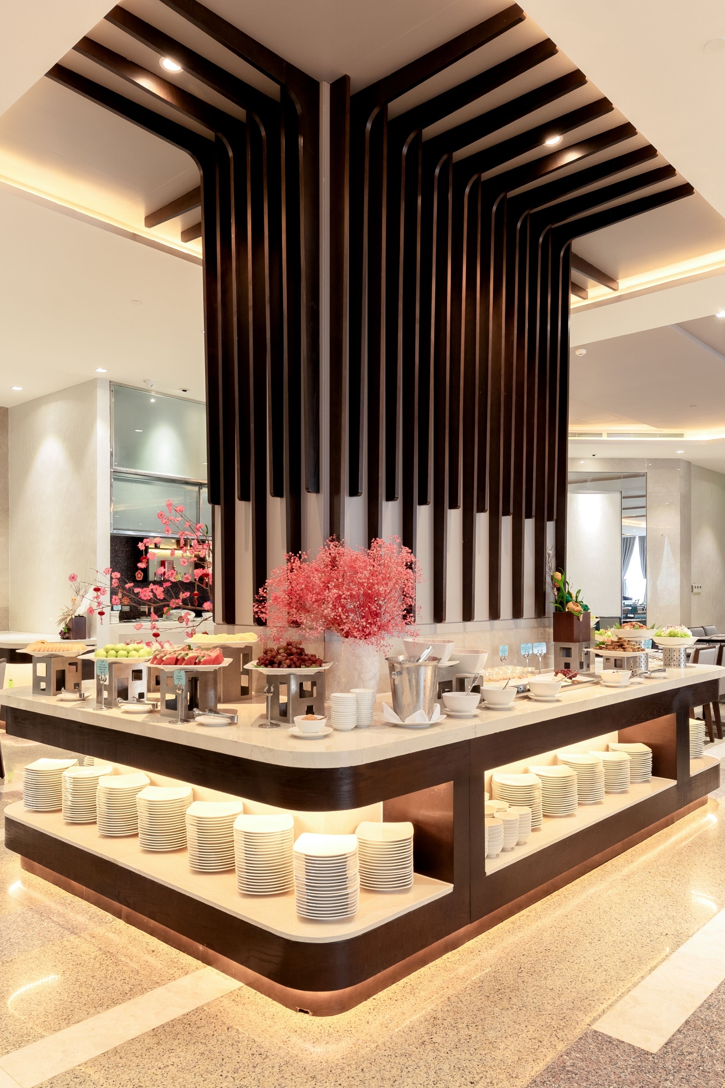 A modern hotel reception area with a central counter displaying food and drink options, a decorative wall with vertical slats, and a clean, polished floor.