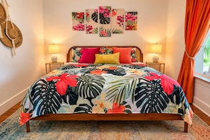 A bed with a floral bedspread, two bedside lamps, and a wall with a tropical-themed artwork. - The Beachcomber – Your Tropical Casa! (Lubbock)