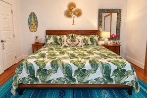 Una cama con estampado de hojas tropicales, cabecera de madera, mesita de noche con lámpara y un adorno en la pared.