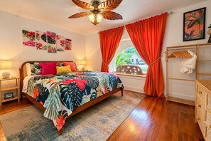 A bedroom with a bed, a window with curtains, a ceiling fan, and a wall with artwork. - The Beachcomber – Your Tropical Casa! (Lubbock)