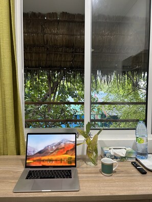 Un ordinateur portable est posé sur un bureau, offrant une vue sur un toit de chaume et la verdure environnante.