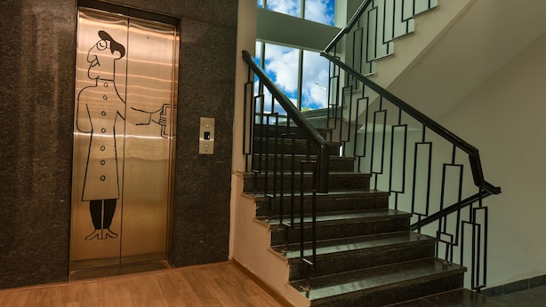 An elevator with a cartoon character, a staircase with black railings, and a view of the sky through the windows. - Hotel Blue Saffron (New Delhi)