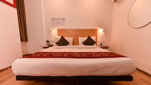 A neatly made bed with a wooden headboard, a red and gold bedspread, and two pillows. There is a nightstand with a lamp and a glass of water. - Hotel Blue Saffron (New Delhi)