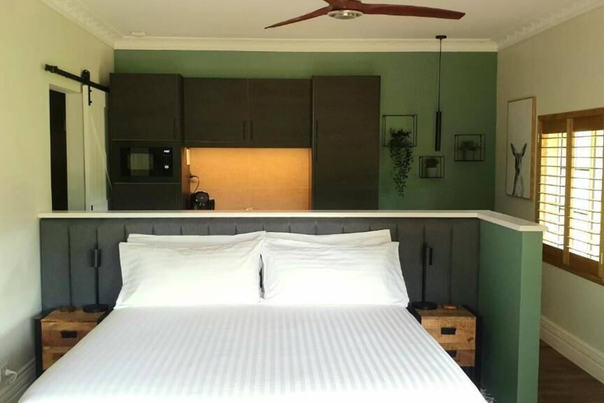Un dormitorio con cama, mesitas de noche de madera, ventilador de techo y vistas a la cocina.