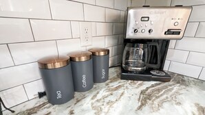 A kitchen with a coffee maker, three grey containers labeled 'Tech', 'Coffee', and 'Sugar', and a tiled backsplash.