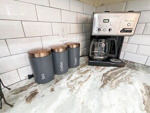 A kitchen with a coffee maker, three grey containers labeled 'Tech', 'Coffee', and 'Sugar', and a tiled backsplash.