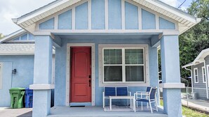 A small house with a red door, a window with blinds, and a porch with a table and chairs.