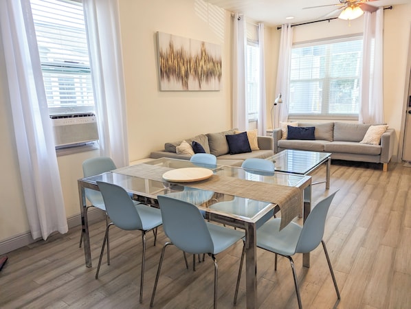 A modern living room with a glass dining table, light blue chairs, a grey sofa, and a large window with white curtains.