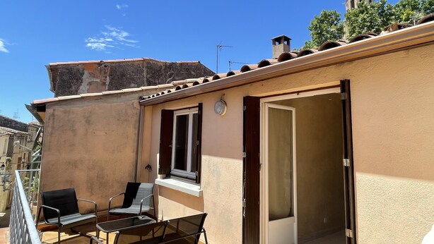 A small outdoor balcony with a table and chairs, a sliding door, and a view of a church tower. - House in the heart of the village (Montagnac)