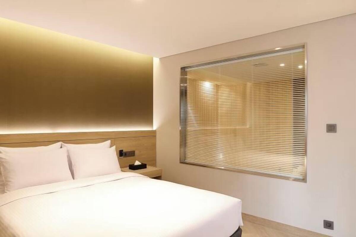 Ein modernes Hotelzimmer mit einem großen Bett, einem Nachttisch, Jalousien und einem großen Fenster.