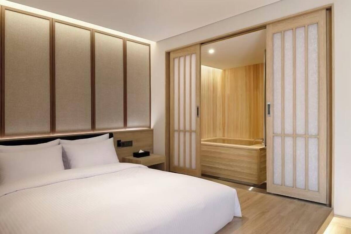 Ein modernes Hotelzimmer mit einem großen Bett, einem Holz-Kopfteil, einem Nachttisch und einer Schiebetür zu einem Badezimmer.