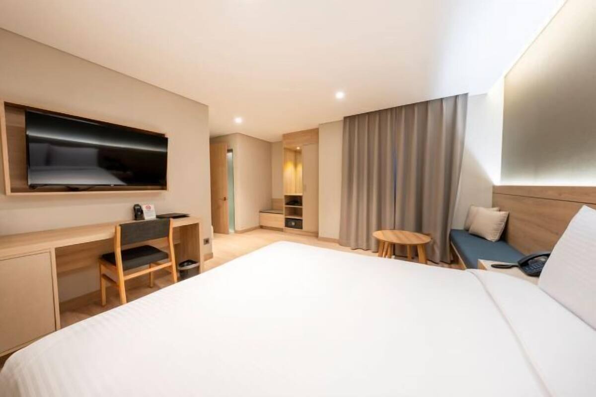 Ein modernes Hotelzimmer mit einem großen Bett, einem Schreibtisch, einem Fernseher, einem kleinen Tisch und einer Sitzecke mit Vorhängen.