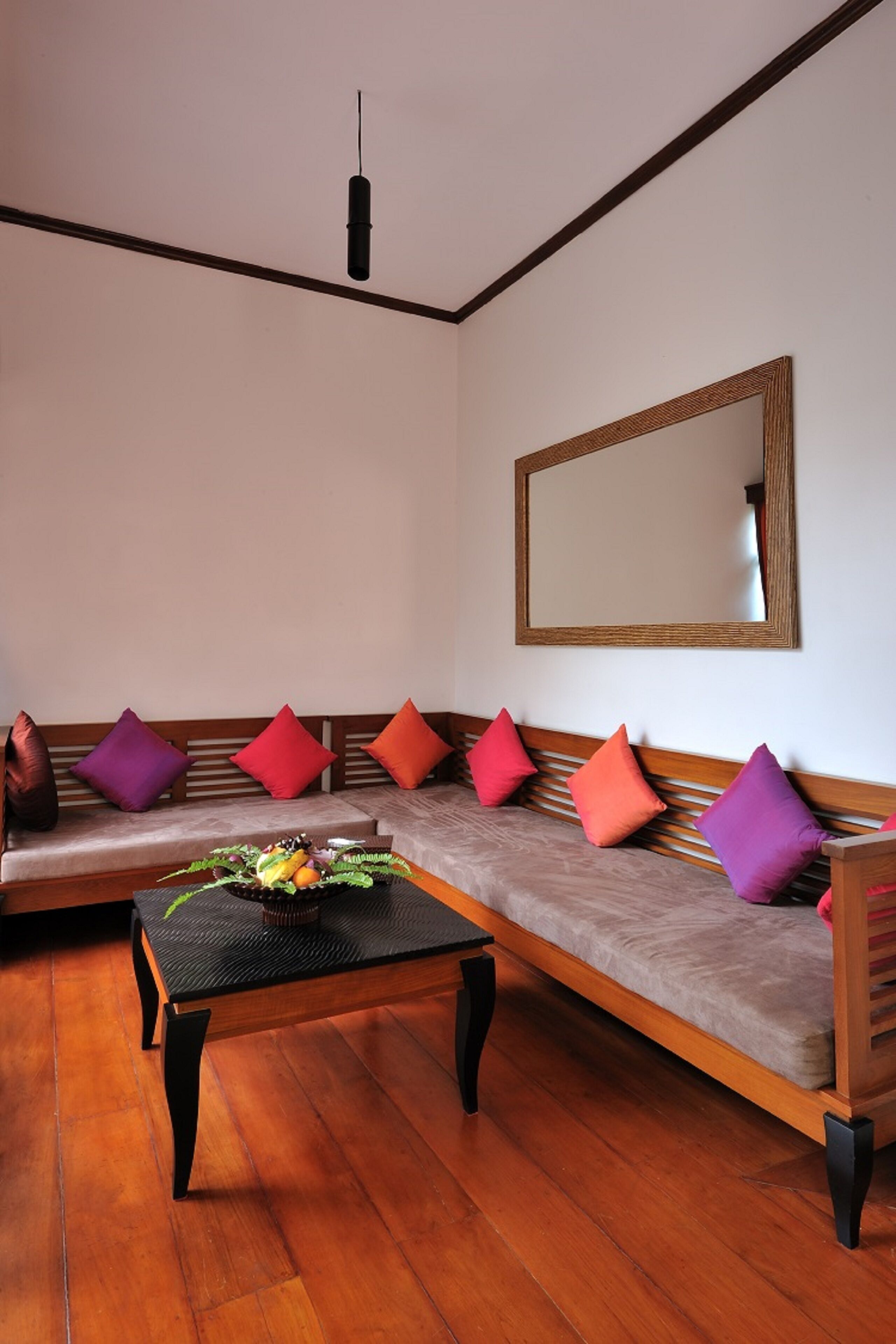 A room with a wooden sofa set against a white wall, a large mirror, and a wooden coffee table with fruit.