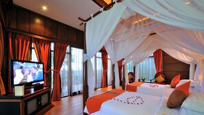 Ein Zimmer mit Fernseher auf einer Holzkommode, zwei Betten mit weißer und orangefarbener Bettwäsche sowie ein Himmelbett.