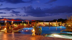 Ein Poolbereich in einer Hotelanlage bei Nacht mit Sonnenschirmen, Liegestühlen und Blick auf die Berge.