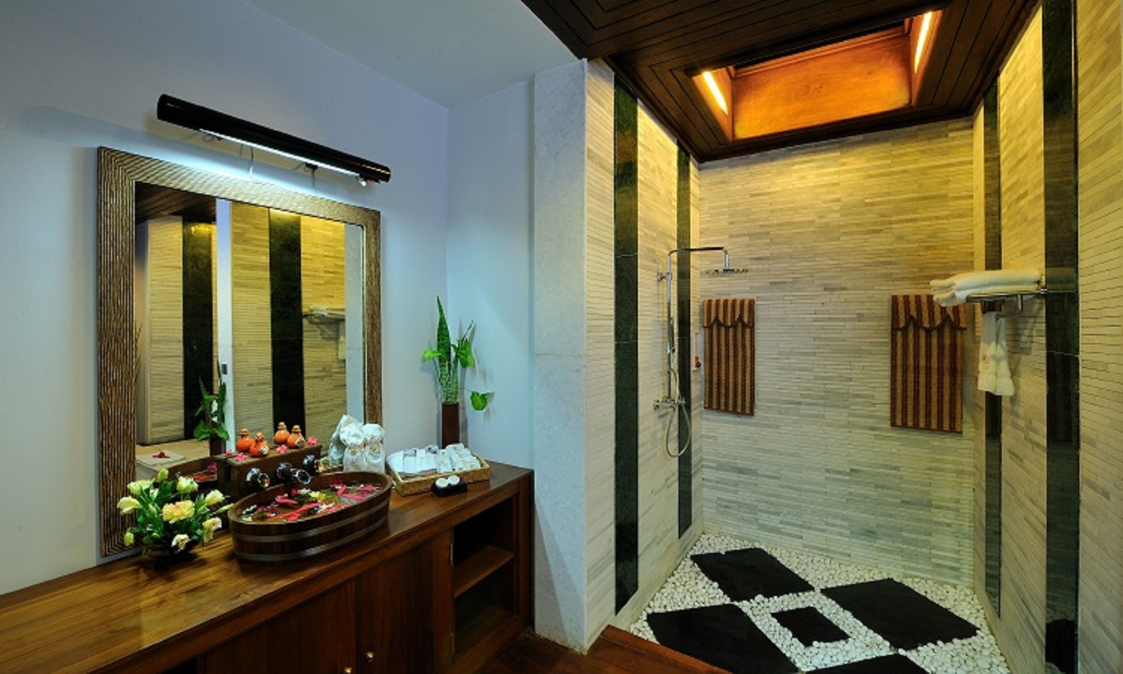 A bathroom with a wooden vanity, a large mirror, a shower with a tiled floor, and a small plant on the vanity.