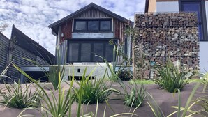 A modern building with a metal facade, a stone wall with stacked firewood, and a landscaped area with tall grasses. - Sleep in the Clouds! The Haiku Hut... (Wentworth Falls)