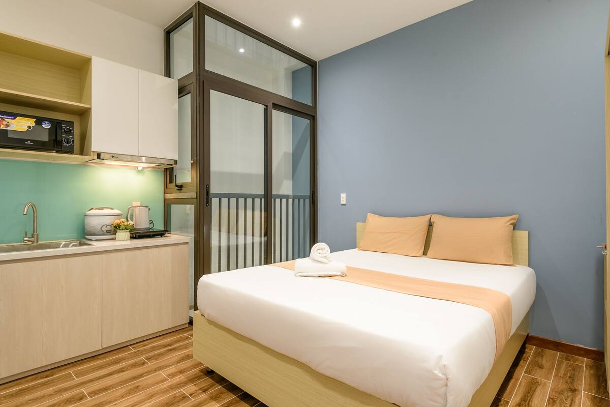 Een compacte hotelkamer met kitchenette, een bed met een beige deken en uitzicht door een groot raam.