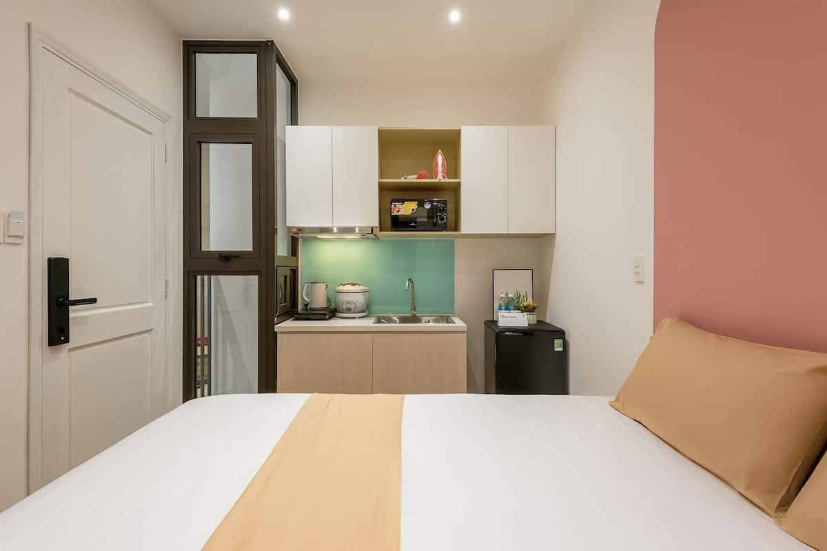 Una habitación de hotel compacta con una cama, una kitchenette, un refrigerador pequeño y una puerta.