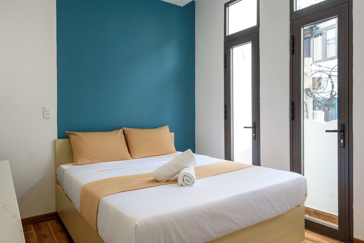 Een net opgemaakt bed met een wit-beige deken, twee beige kussens en een opgerolde handdoek op een houten hoofdbord in een kamer met teal muren en een groot raam.