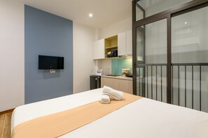 Habitación de hotel moderna con una cama, televisor de pantalla plana montado en la pared, kitchenette con lavamanos y encimera, y puerta corrediza de vidrio que da a un balcón.
