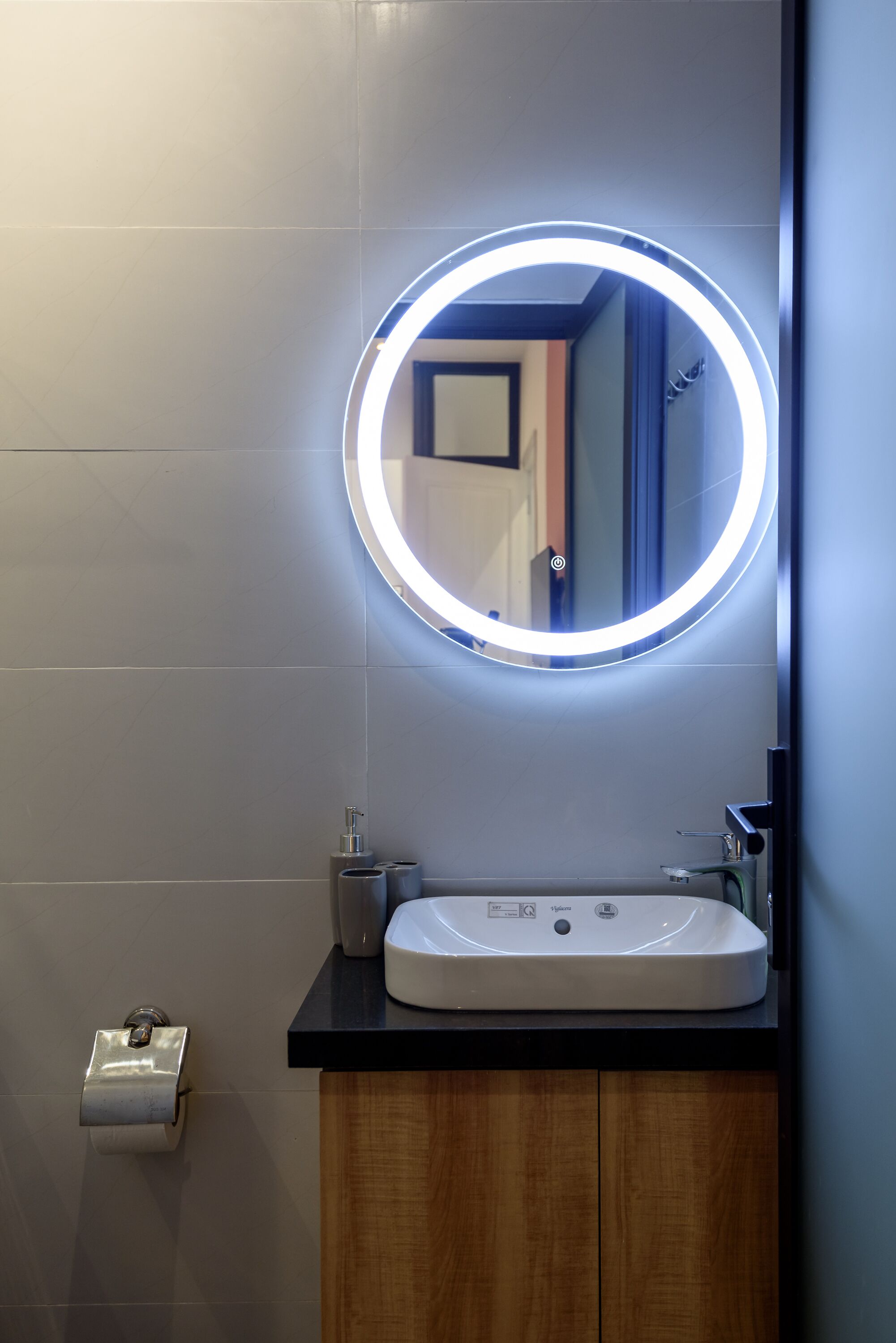 A modern bathroom with a round mirror, illuminated by a circular light, a white sink, and a wooden cabinet.