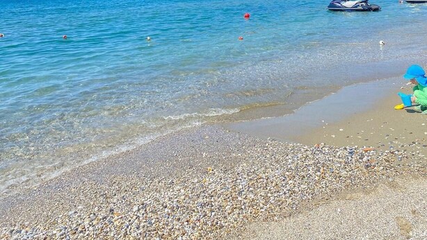 A pebble beach with clear blue water, boats, and a distant hill with buildings. - Respect Apart Hotel (Alanya)