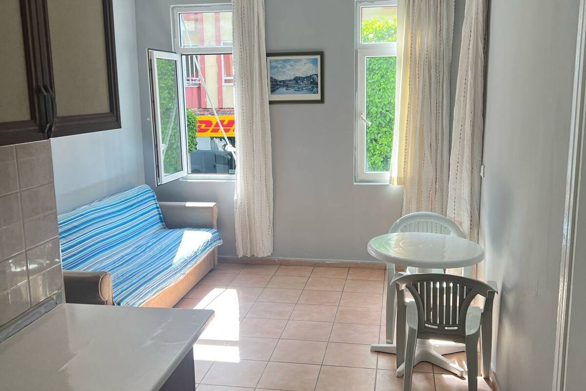 A compact kitchen with a sink and countertop, a small dining area with a table and chairs, a sofa with a striped cushion, and a window with curtains.