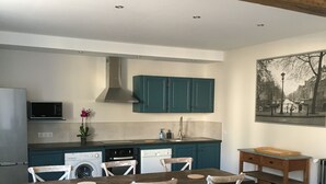 A modern kitchen with teal cabinets, a dining area with a wooden table and white chairs, and a large dark green sofa in the foreground. - Gîte "La Hulotte" country property away from the city (Villeneuve-la-Lionne)