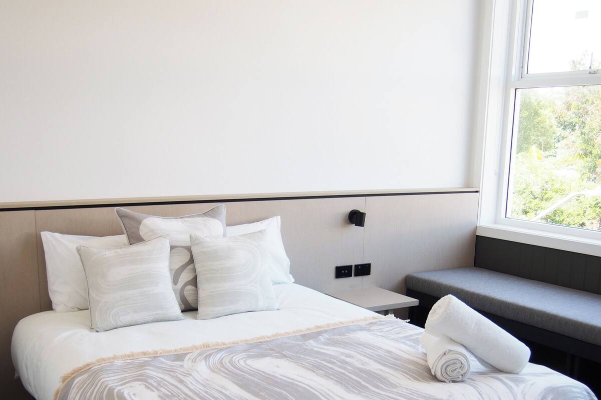 Una cama bien tendida con una manta gris y blanca de estampado, una mesita de noche y una ventana con vistas a la vegetación.