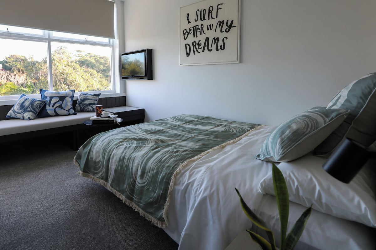 Un dormitorio con cama, un rincón con cojines junto a la ventana, un cuadro colgado en la pared y una cita motivacional.
