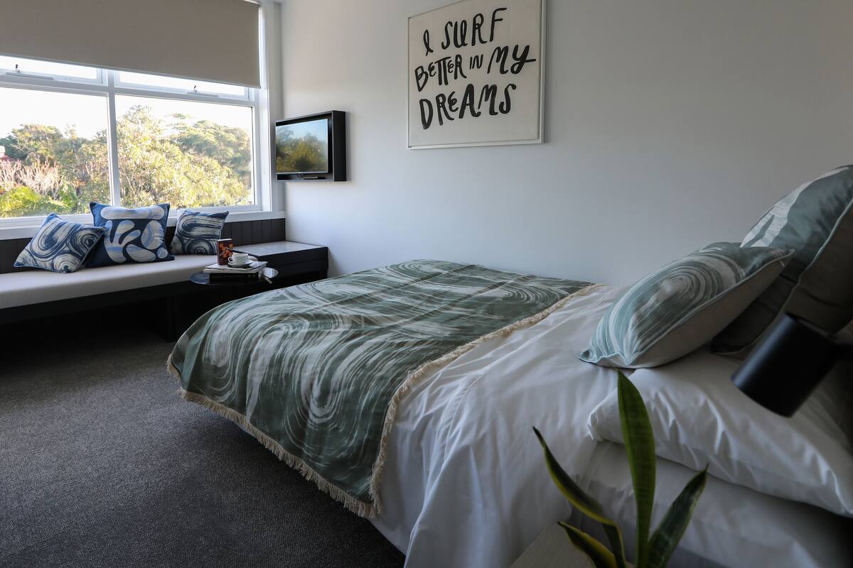 Un dormitorio con cama, un rincón con cojines junto a la ventana, un cuadro colgado en la pared y una cita motivacional.