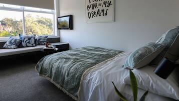 Une chambre à coucher avec un lit, un coin fenêtre agrémenté de coussins, un tableau accroché au mur et une citation inspirante.