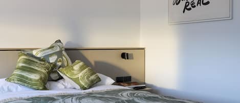 A neatly made bed with a patterned bedspread, pillows, and a bedside table with a book and a phone. A wall-mounted light and a sign with text are also visible.