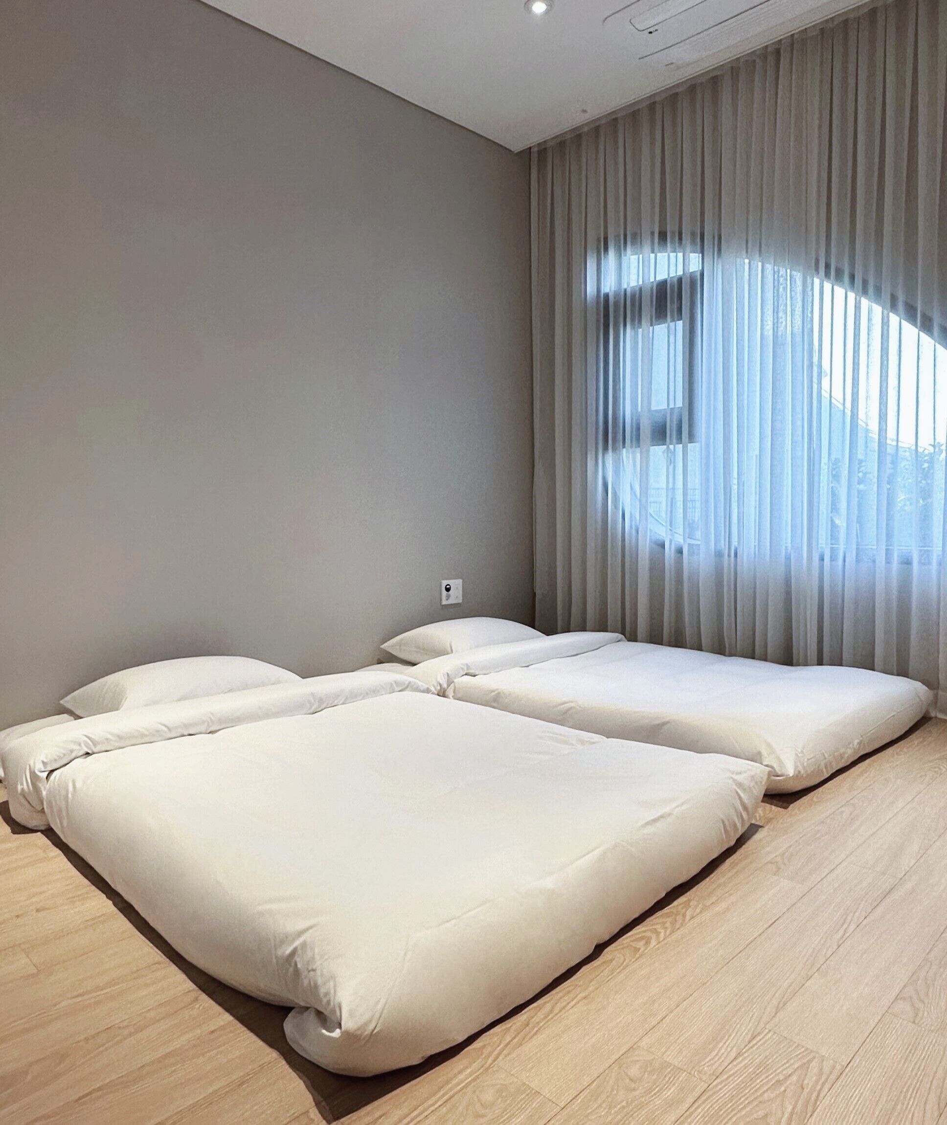 Una habitación de hotel moderna con dos camas, un ventanal con cortinas translúcidas y piso de madera clara.
