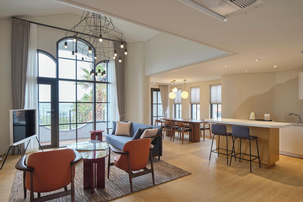 A modern hotel room with a sofa, dining table, chairs, and a kitchen area.