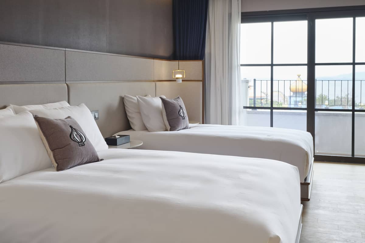 A hotel room with two beds, a nightstand, a lamp, and a large window with a view.