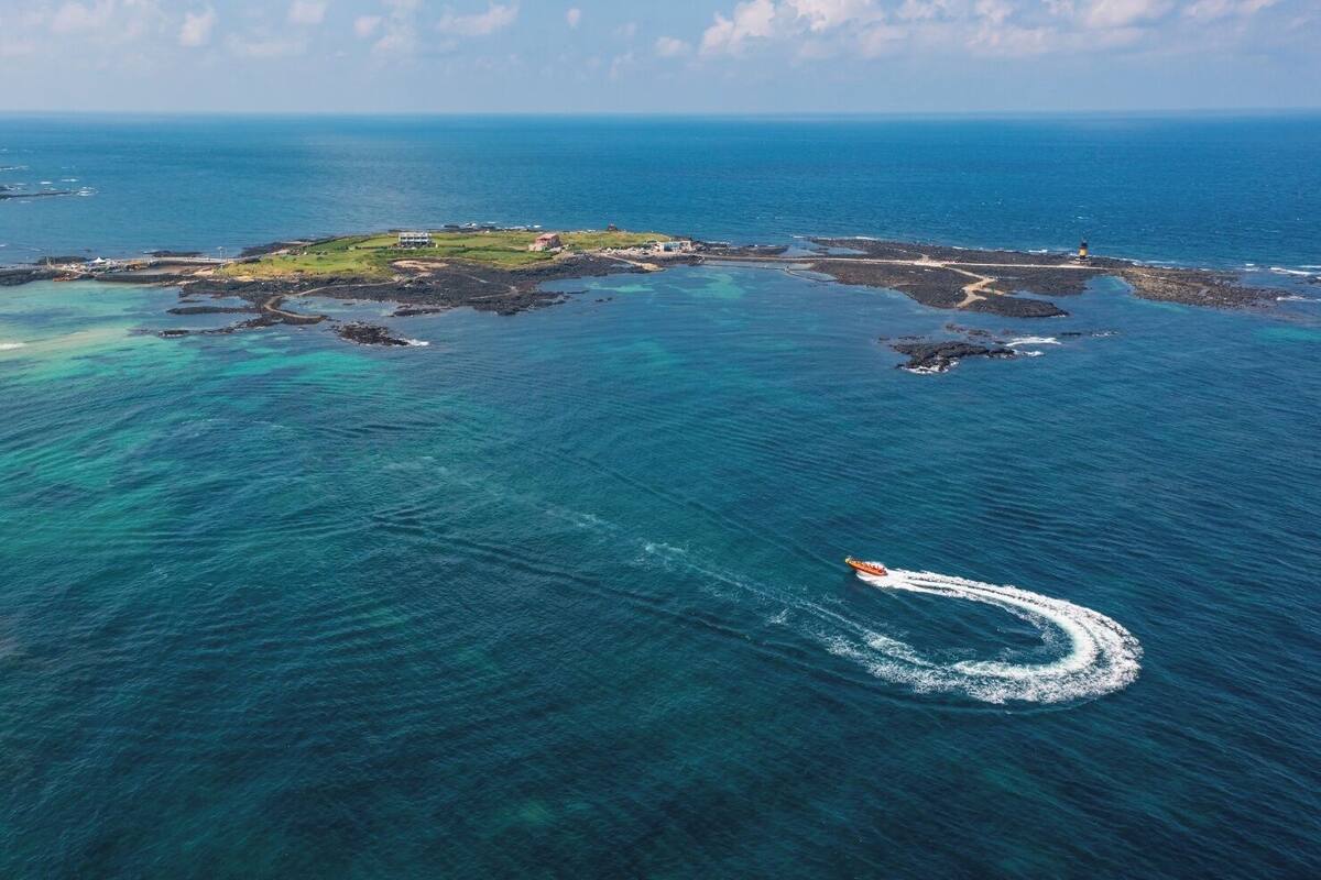 An aerial view of a coastal area with clear blue waters, a small island with buildings, and a lighthouse.
