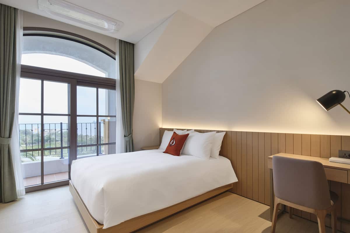 A modern hotel room with a large bed, a desk, and a view of the outdoors through a large window.