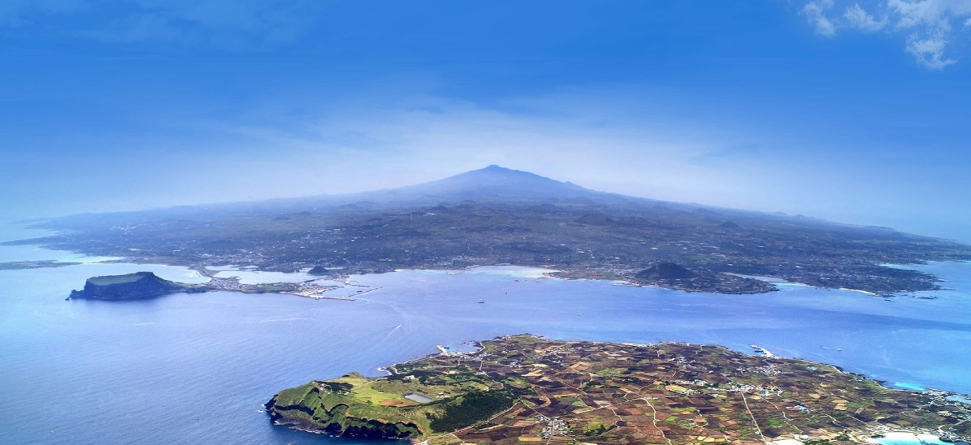 An aerial view of a coastal area with a prominent mountain in the background, a small island with visible buildings, and a large body of water.