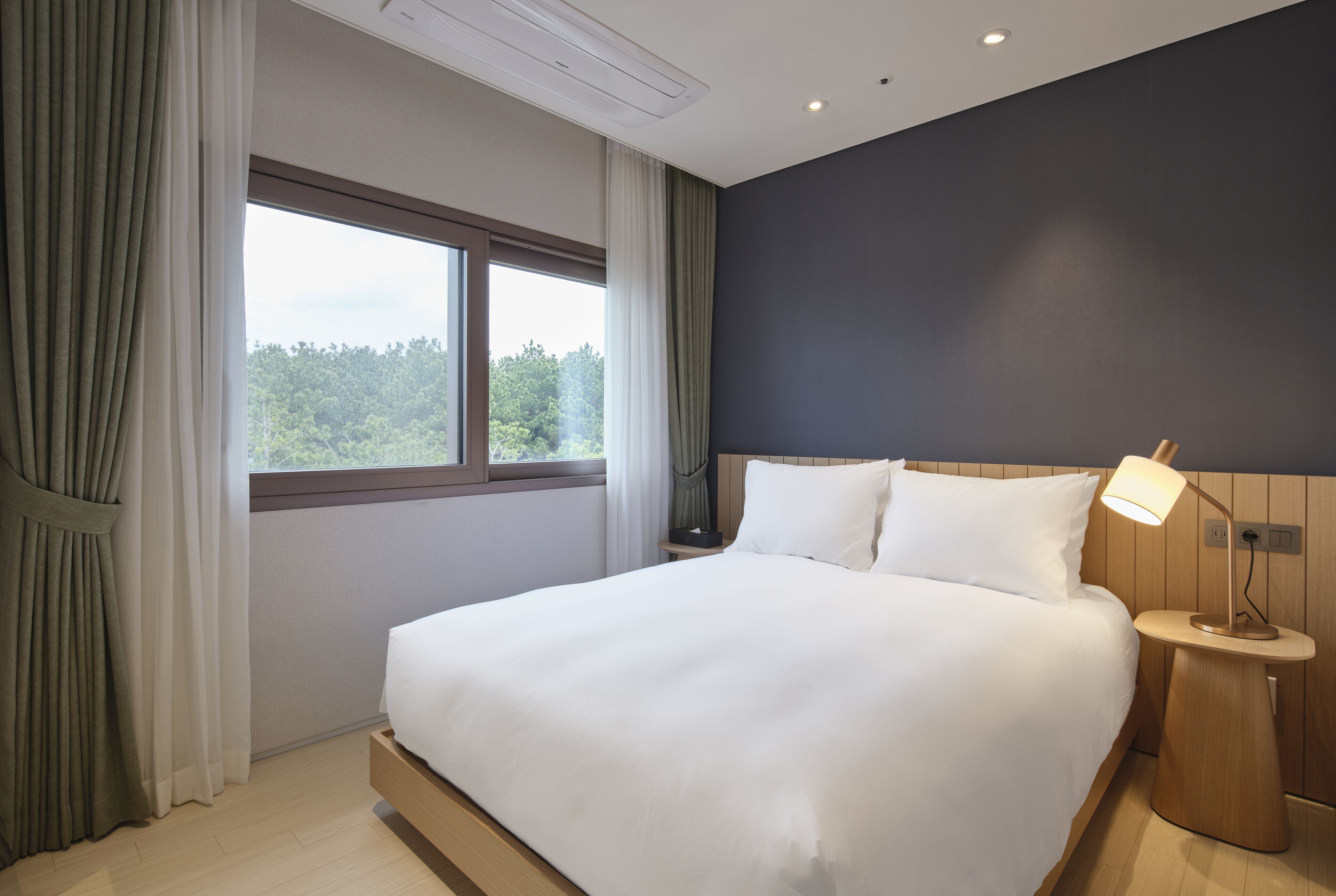 A modern hotel room with a large bed, a nightstand, a lamp, and a window with curtains.