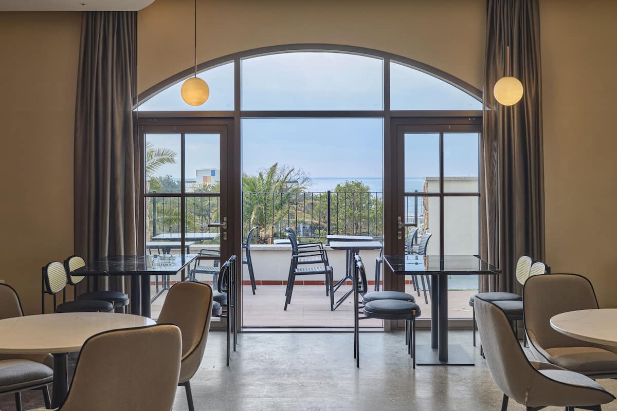 A modern dining area with glass tables and chairs, large windows with curtains, and a view of the sea.