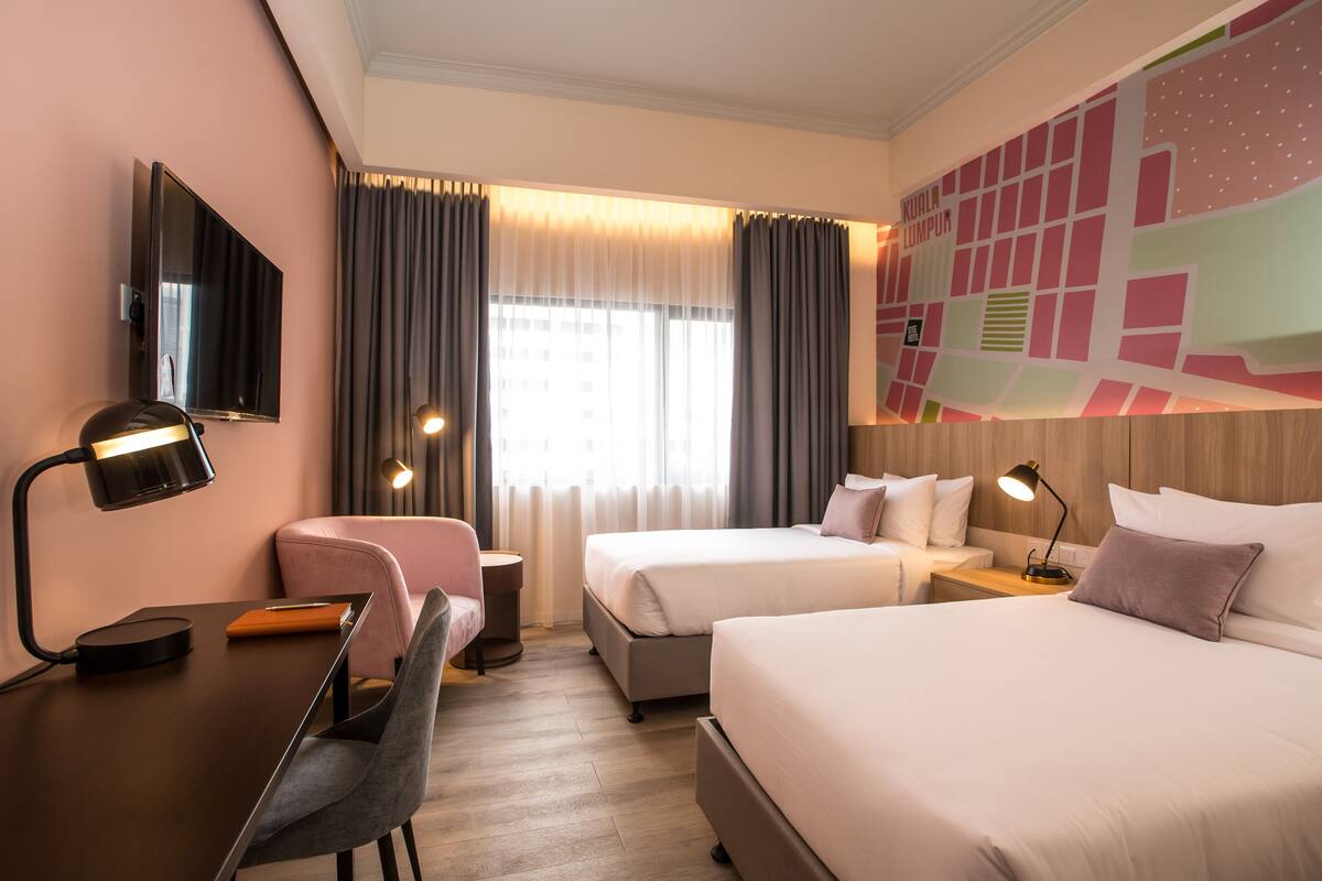 A hotel room with two beds, a desk with a chair, a TV, and a cityscape mural on the wall.