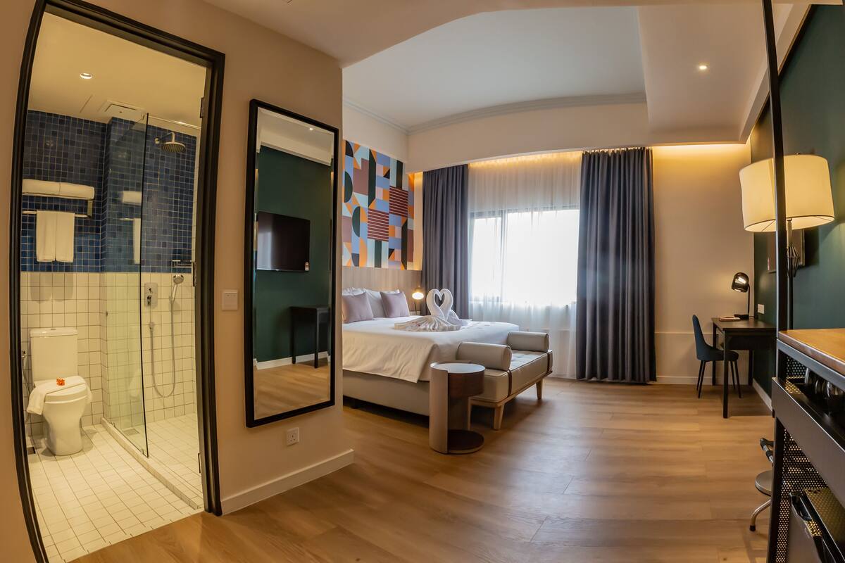 A modern hotel room with a bed, bedside table, desk, chair, and a shower area.