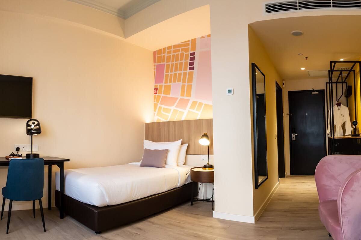 A hotel room with a bed, desk, chair, and a pink armchair.